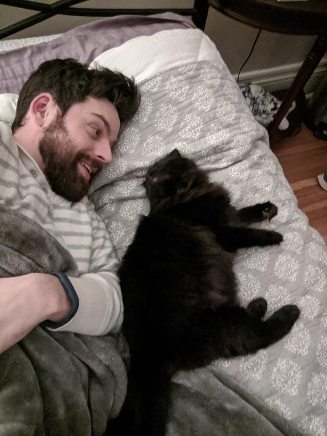 Me and Taggy relaxing on a bed, with Taggy to her right. Her back is towards me, but she turns her head to look me in the eyes while I smile.