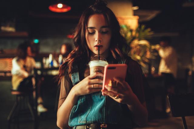 A teenage girl in a coffee shop looks at her phone while sipping a drink