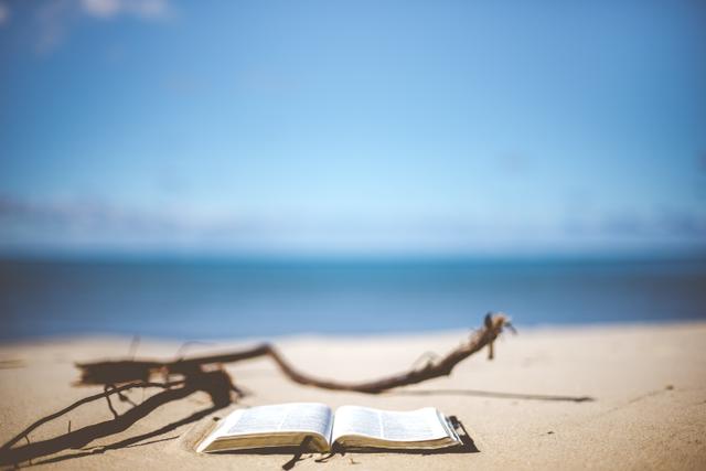 An open book lying on a sandy beach, with the water and sky visible in the background. No people are pictured nearby.