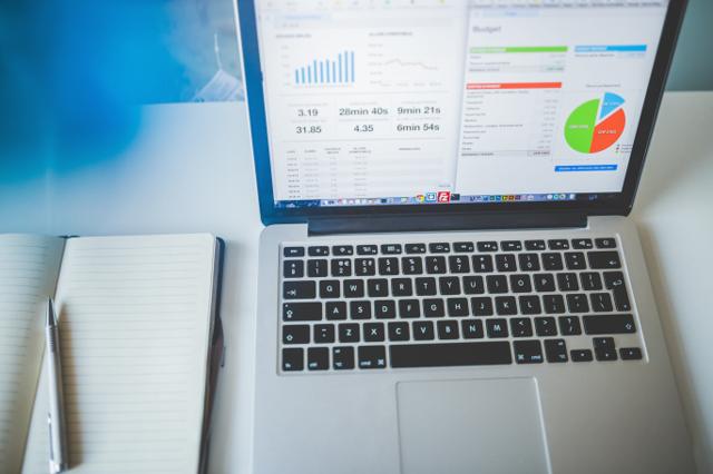 An Apple laptop shows various charts, graphs, and other metrics, while a physical notebook and a pen rest to its left